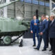 Group of officials in suits examining a large green armored tank on display in a bright hangar-like showroom.