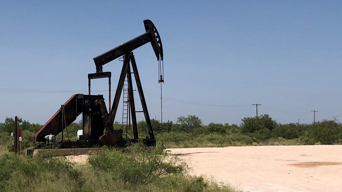 Oil pump jack in a dry, grassy field under a clear blue sky, with distant shrubs and utility poles nearby
