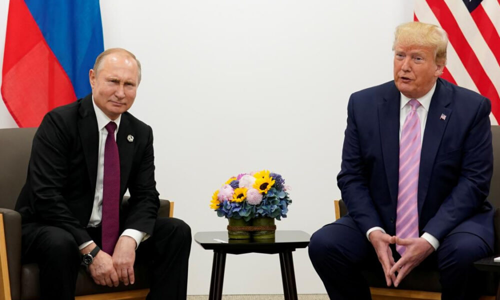 Two men in dark suits sit across from each other in a formal meeting, with a bouquet of flowers on the table and national flags in the background.
