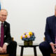 Two men in dark suits sit across from each other in a formal meeting, with a bouquet of flowers on the table and national flags in the background.