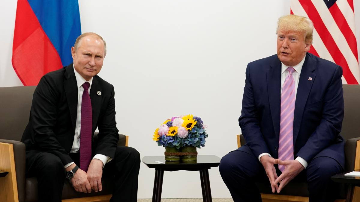 Two men in dark suits sit across from each other in a formal meeting, with a bouquet of flowers on the table and national flags in the background.
