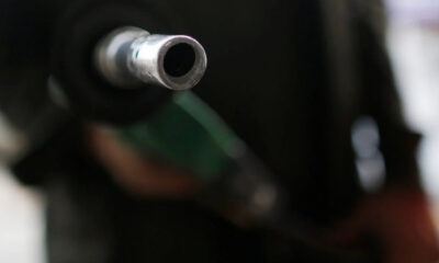 Metal fuel nozzle pointed toward the camera at a gas station, with a blurred background of a person holding the hose