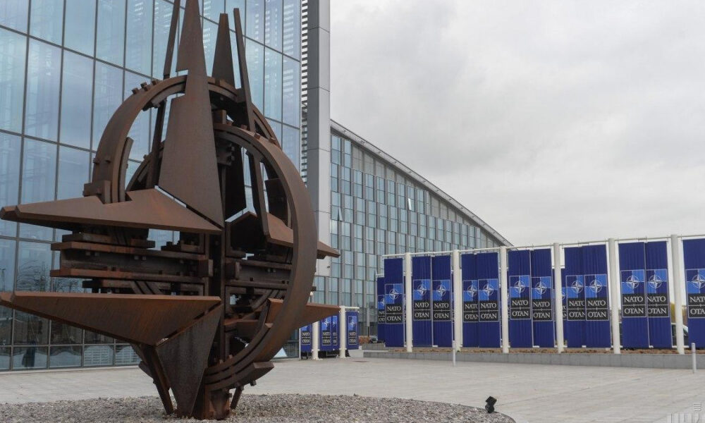 Large rust-colored metal sculpture with circular rings and triangular blades outside a glass-fronted building; blue NATO banners line the facade in the background.