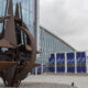 Large rust-colored metal sculpture with circular rings and triangular blades outside a glass-fronted building; blue NATO banners line the facade in the background.