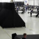 Spacious factory floor with long rows of black fabric panels and workers at workbenches assembling components.
