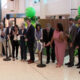 Group of diverse professionals at a ribbon-cutting ceremony with green balloons indoors in a mall or atrium.