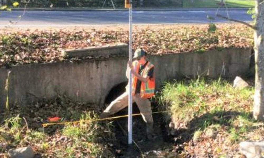 Construction worker in a reflective vest stands by a drainage culvert, holding a measuring pole for inspection.