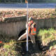 Construction worker in a reflective vest stands by a drainage culvert, holding a measuring pole for inspection.