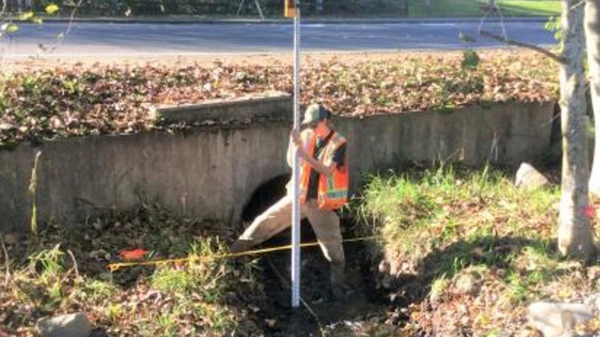 Construction worker in a reflective vest stands by a drainage culvert, holding a measuring pole for inspection.