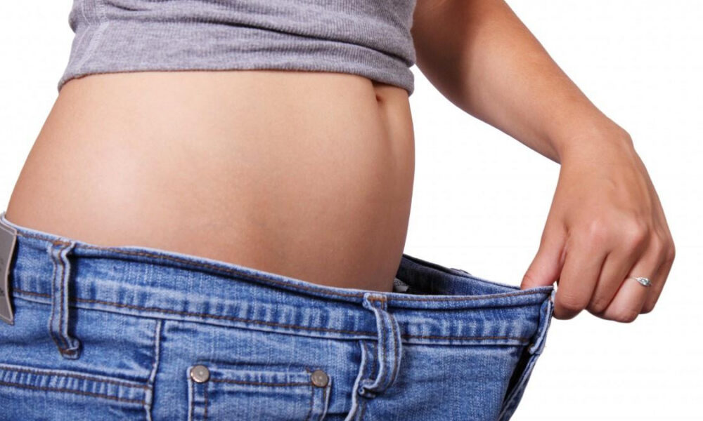 Person's midsection with a rounded belly as they pull the waistband of loose jeans away, illustrating weight loss or a loose fit, side view