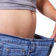 Person's midsection with a rounded belly as they pull the waistband of loose jeans away, illustrating weight loss or a loose fit, side view