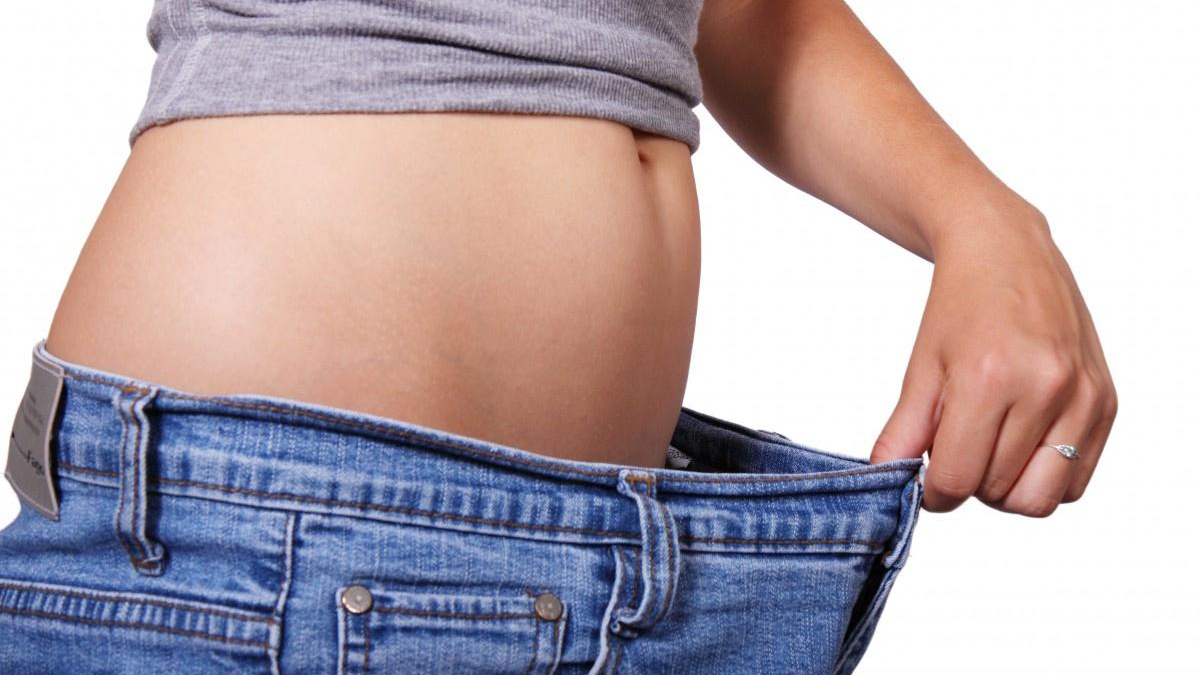 Person's midsection with a rounded belly as they pull the waistband of loose jeans away, illustrating weight loss or a loose fit, side view