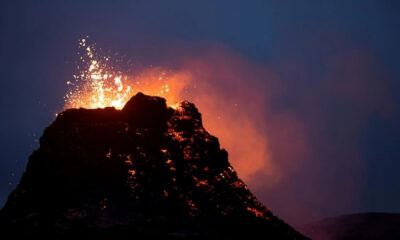 Volcano eruption at night with molten lava glowing on dark rock and sparks shooting upward from the summit.