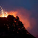 Volcano eruption at night with molten lava glowing on dark rock and sparks shooting upward from the summit.