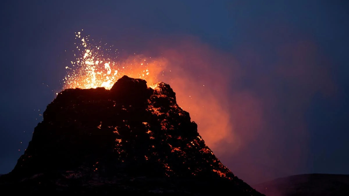 Volcano eruption at night with molten lava glowing on dark rock and sparks shooting upward from the summit.