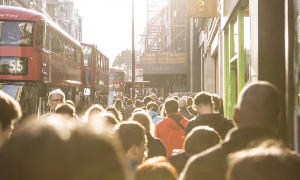 Crowded city street with red double-decker buses and pedestrians in sunlight near a shopfront (Primark).