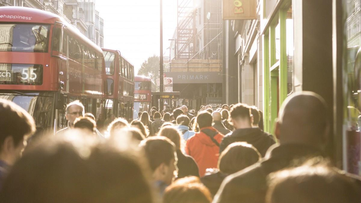 Crowded city street with red double-decker buses and pedestrians in sunlight near a shopfront (Primark).