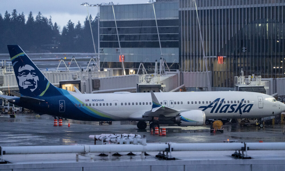Alaska Airlines aircraft parked at a gate on a wet tarmac with jet bridges and a modern terminal building in the background.
