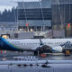 Alaska Airlines aircraft parked at a gate on a wet tarmac with jet bridges and a modern terminal building in the background.