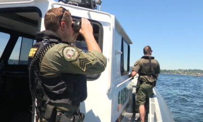 Two uniformed officers on a white patrol boat, one using binoculars to scan the water while the other stands at the side ready on a sunny day by the shoreline.