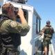 Two uniformed officers on a white patrol boat, one using binoculars to scan the water while the other stands at the side ready on a sunny day by the shoreline.