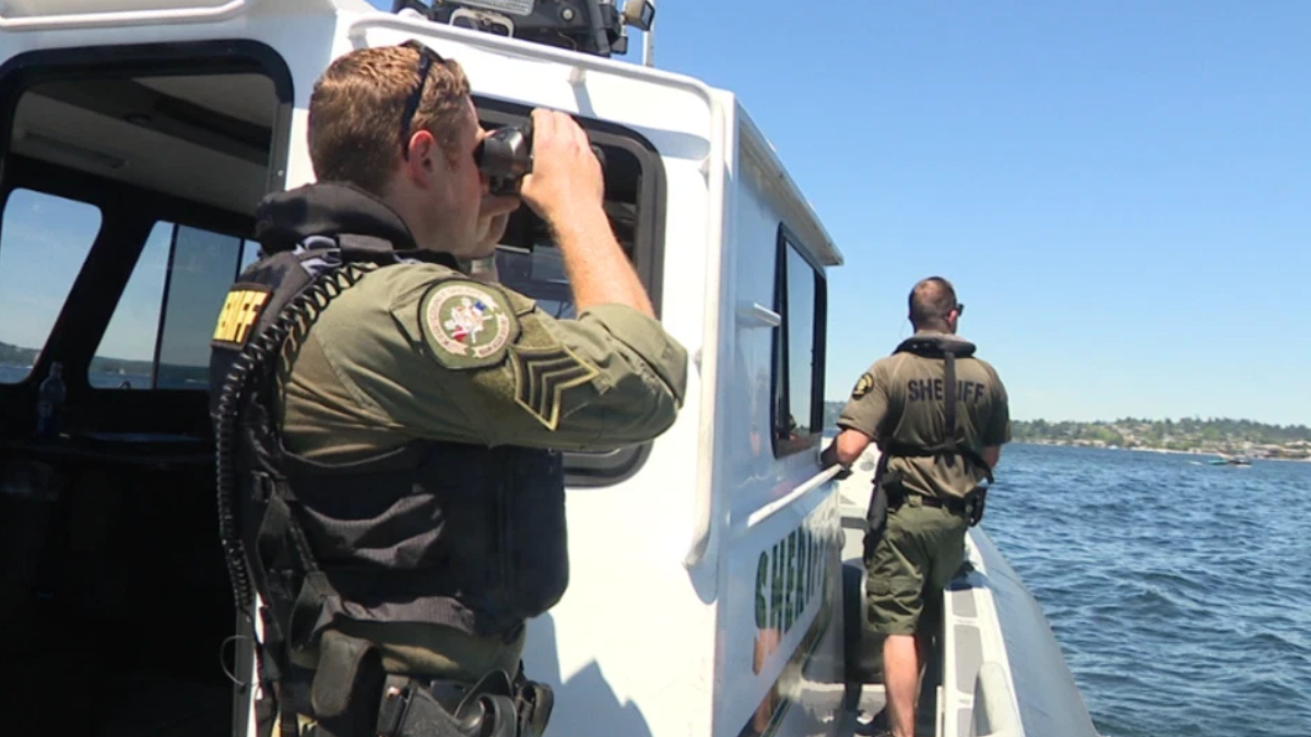 Two uniformed officers on a white patrol boat, one using binoculars to scan the water while the other stands at the side ready on a sunny day by the shoreline.