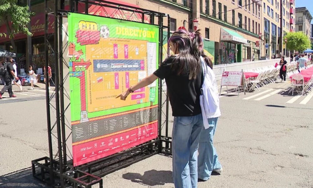 Two people read a large colorful event map mounted on a metal frame at a sunny street festival with chairs and storefronts in the background.