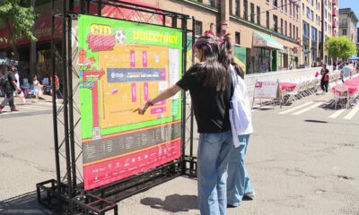 Two people read a large colorful event map mounted on a metal frame at a sunny street festival with chairs and storefronts in the background.