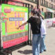 Two people read a large colorful event map mounted on a metal frame at a sunny street festival with chairs and storefronts in the background.
