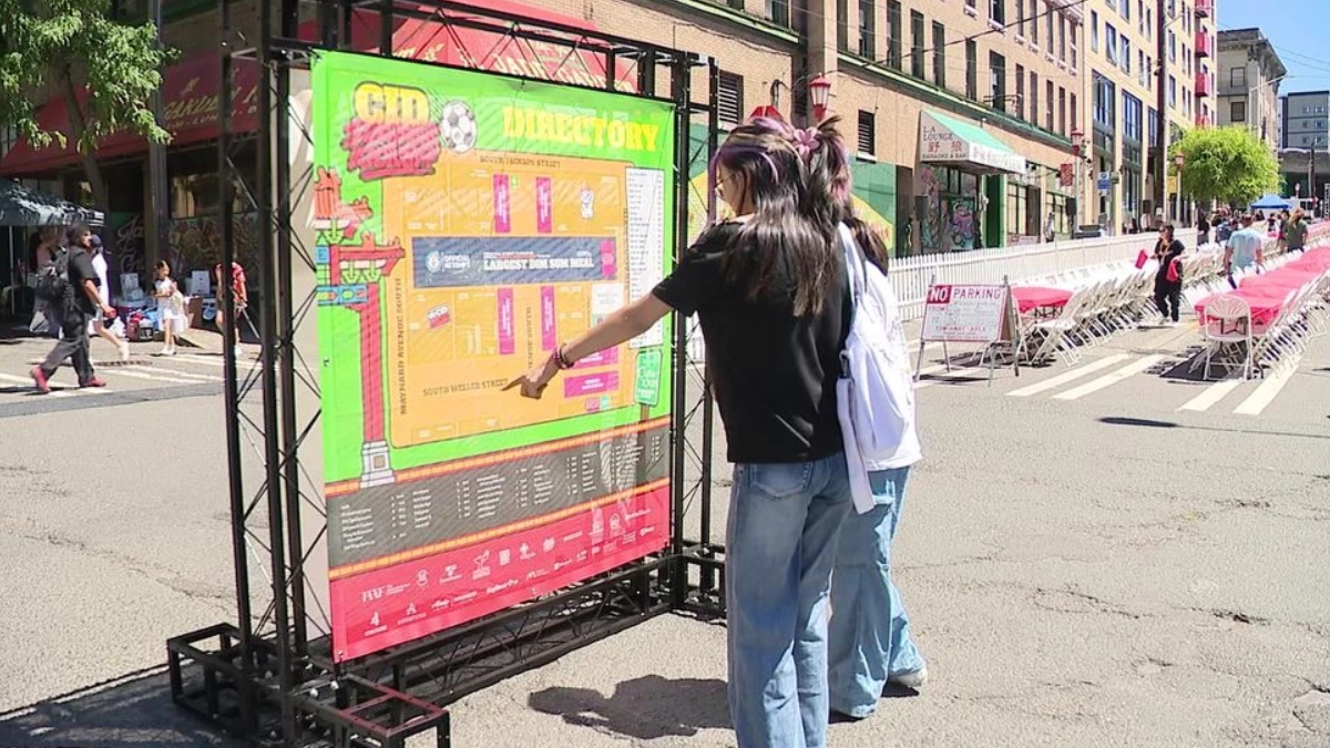 Two people read a large colorful event map mounted on a metal frame at a sunny street festival with chairs and storefronts in the background.