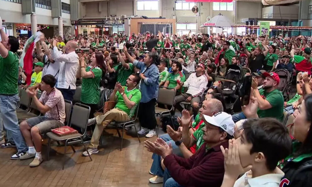 Large crowd of people seated and standing in an indoor venue, many wearing green, cheering and clapping at an event.