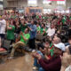 Large crowd of people seated and standing in an indoor venue, many wearing green, cheering and clapping at an event.
