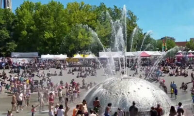 Crowd gathers around a large silver fountain dome with water jets shooting upward at an outdoor festival. Details: tents line the background on a sunny day.