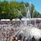 Crowd gathers around a large silver fountain dome with water jets shooting upward at an outdoor festival. Details: tents line the background on a sunny day.