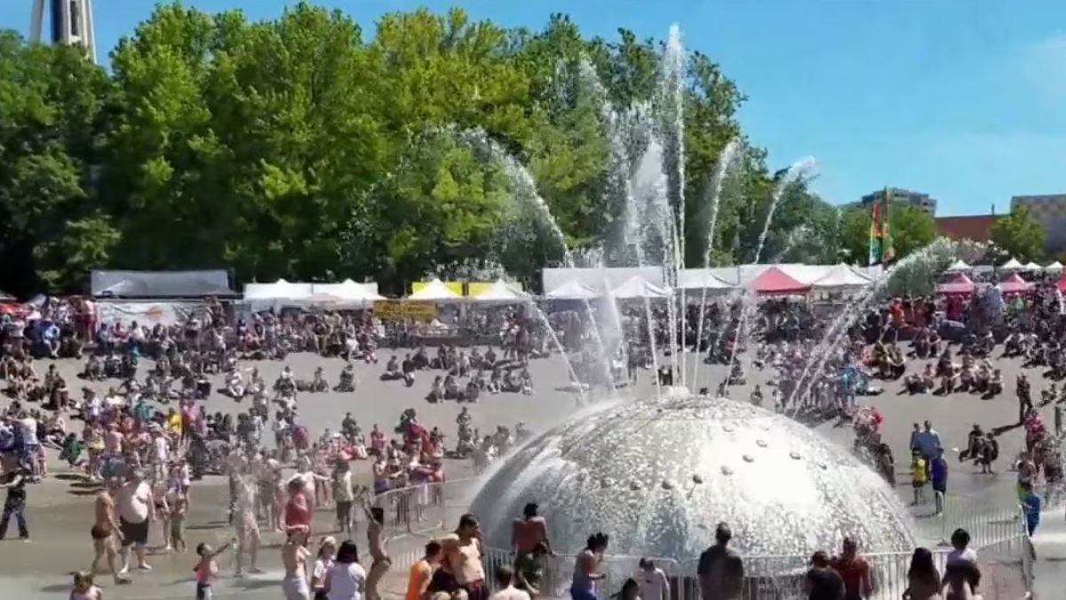 Crowd gathers around a large silver fountain dome with water jets shooting upward at an outdoor festival. Details: tents line the background on a sunny day.