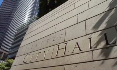 Stone wall with carved letters spelling 'Seattle City Hall' as tall office buildings loom in the background.