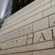 Stone wall with carved letters spelling 'Seattle City Hall' as tall office buildings loom in the background.