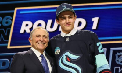 Two men posing and smiling on a stage, one in a suit and the other in a dark hockey jersey, with a ROUND 1 sign in the background.