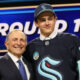 Two men posing and smiling on a stage, one in a suit and the other in a dark hockey jersey, with a ROUND 1 sign in the background.