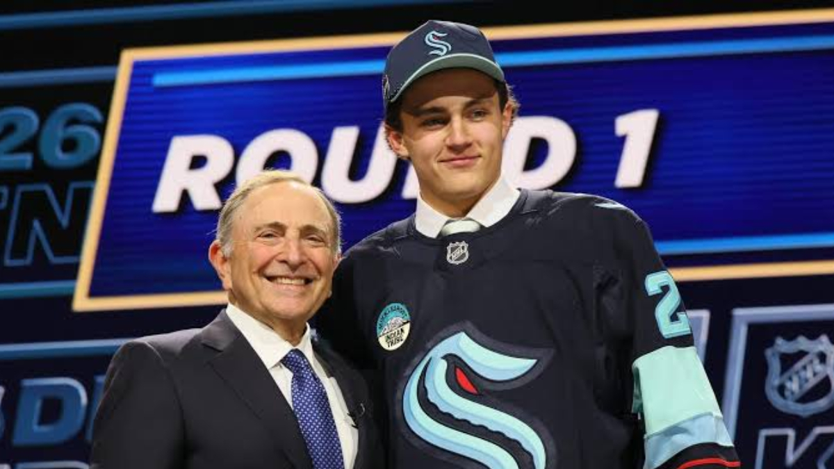 Two men posing and smiling on a stage, one in a suit and the other in a dark hockey jersey, with a ROUND 1 sign in the background.