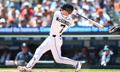 Baseball batter in a white uniform swinging a bat during a game with a blurred crowd in the background.