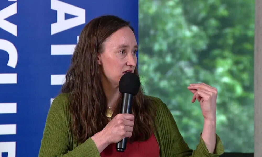 Panelist in a green cardigan speaks into a handheld microphone, gesturing with her right hand on stage with a blue banner behind her.