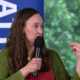 Panelist in a green cardigan speaks into a handheld microphone, gesturing with her right hand on stage with a blue banner behind her.