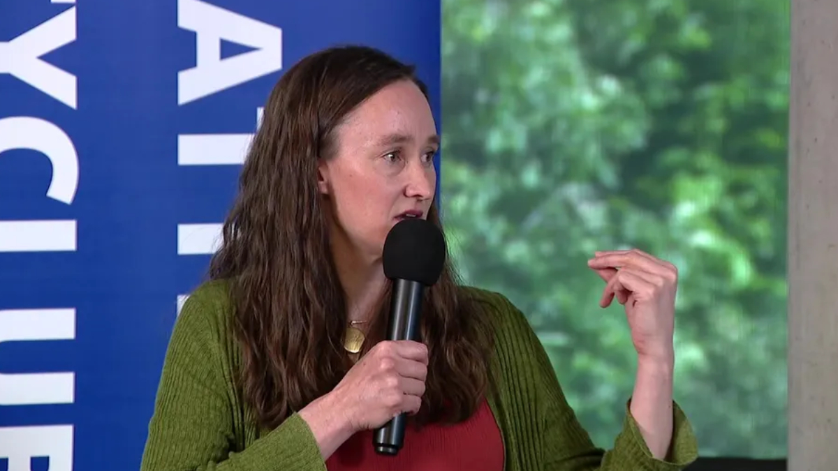 Panelist in a green cardigan speaks into a handheld microphone, gesturing with her right hand on stage with a blue banner behind her.