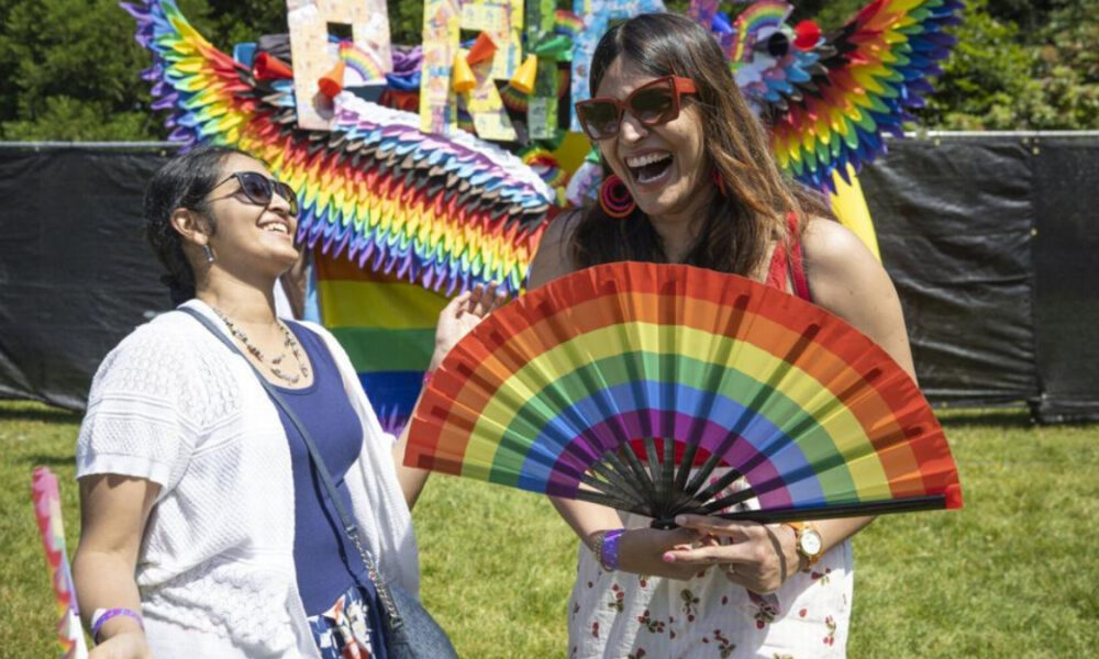 Two women laugh together at an outdoor Pride festival, one holding a bright rainbow fan in front of her.