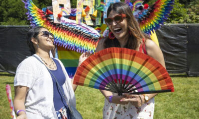Two women laugh together at an outdoor Pride festival, one holding a bright rainbow fan in front of her.