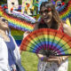 Two women laugh together at an outdoor Pride festival, one holding a bright rainbow fan in front of her.