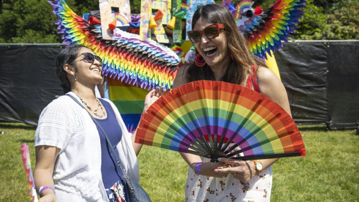 Two women laugh together at an outdoor Pride festival, one holding a bright rainbow fan in front of her.