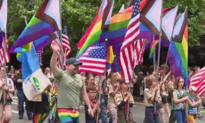 Parade on a street with marchers carrying rainbow Pride flags and U.S. flags, celebrating together.