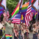Parade on a street with marchers carrying rainbow Pride flags and U.S. flags, celebrating together.